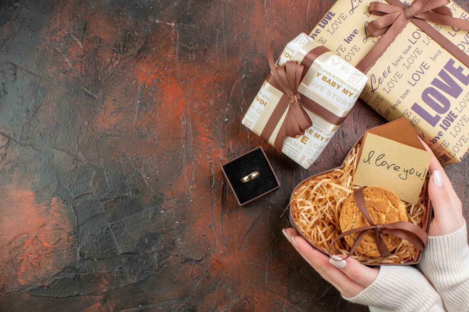 Top view of Valentine’s Day gift boxes with an engagement ring, cookies, and love note on a dark textured background, symbolising romance and commitment.