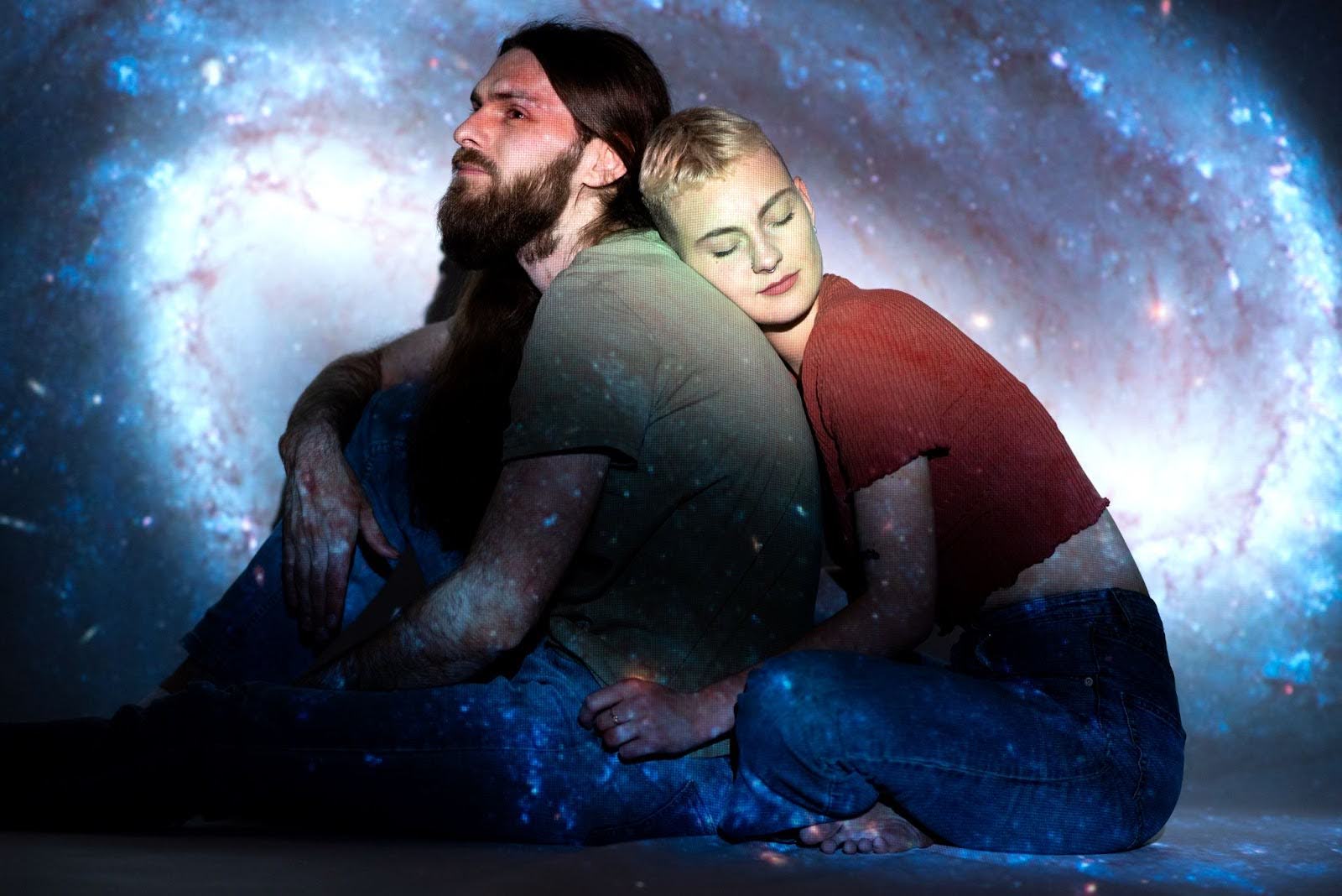 Man and woman sitting back to back with a galaxy projection, symbolising emotional intimacy, spiritual connection, and cosmic love.